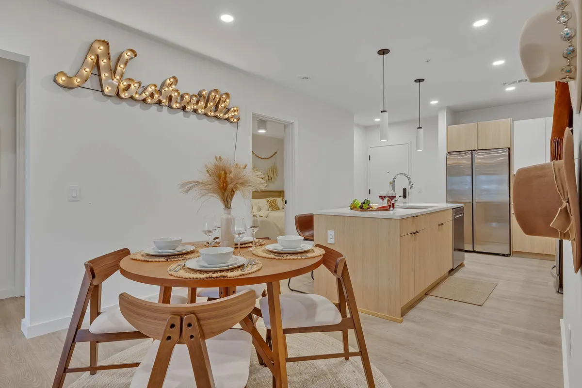 A warm and welcoming dining nook set for four, complete with plates and glasses, sits beside a kitchen island with stools. The decor includes a playful Nashville sign and soft lighting, enhancing the hospitable atmosphere. The view balances both the dining and kitchen preparation spaces, evoking the spirit of shared meals and celebrations.