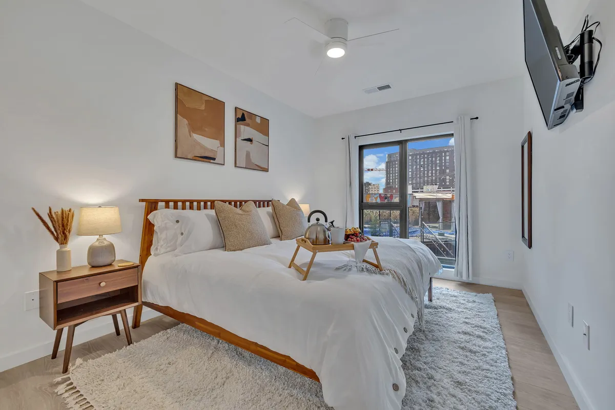 A serene bedroom with a plush bed, large window offering natural light, decorative artwork above the headboard, a nightstand with a lamp, and a breakfast tray set atop the bedding. The room exudes a cozy, restful vibe and appears designed for privacy and deep relaxation.