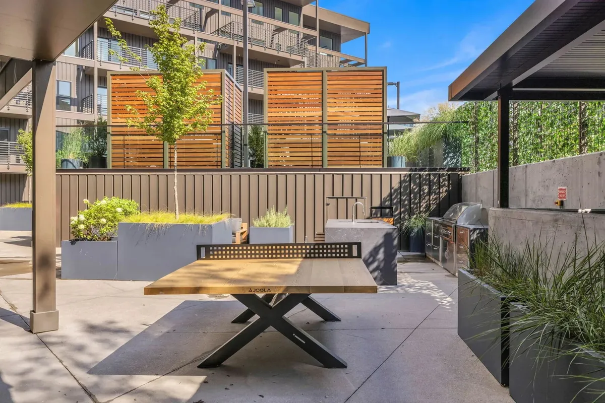 An inviting outdoor grill and dining area with a wooden table, lush green plants, and wooden privacy screens. The space is perfect for group barbecues or relaxed outdoor meals, with a comfortable, social vibe.