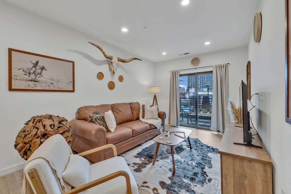 A cozy living room with a leather sofa, decorative wall art, a cowhide rug, and a wooden coffee table. Large windows bathe the space in natural light, creating an inviting setting for unwinding or socializing.