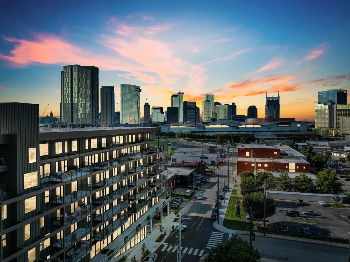 This photo features a modern apartment building with the Nashville city skyline and a beautiful sunset sky in the background. The image captures the dynamic urban environment and highlights the property’s proximity to downtown offices and vibrant city life.