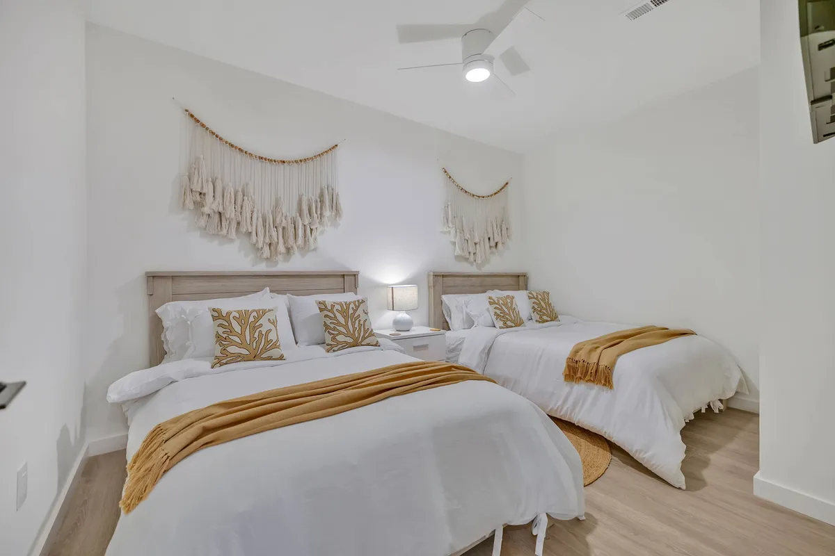 A spacious bedroom featuring two large beds with decorative pillows, textured wall hangings, and nightstands with lamps on either side. The atmosphere is calm and welcoming, ideal for accommodating groups or families.