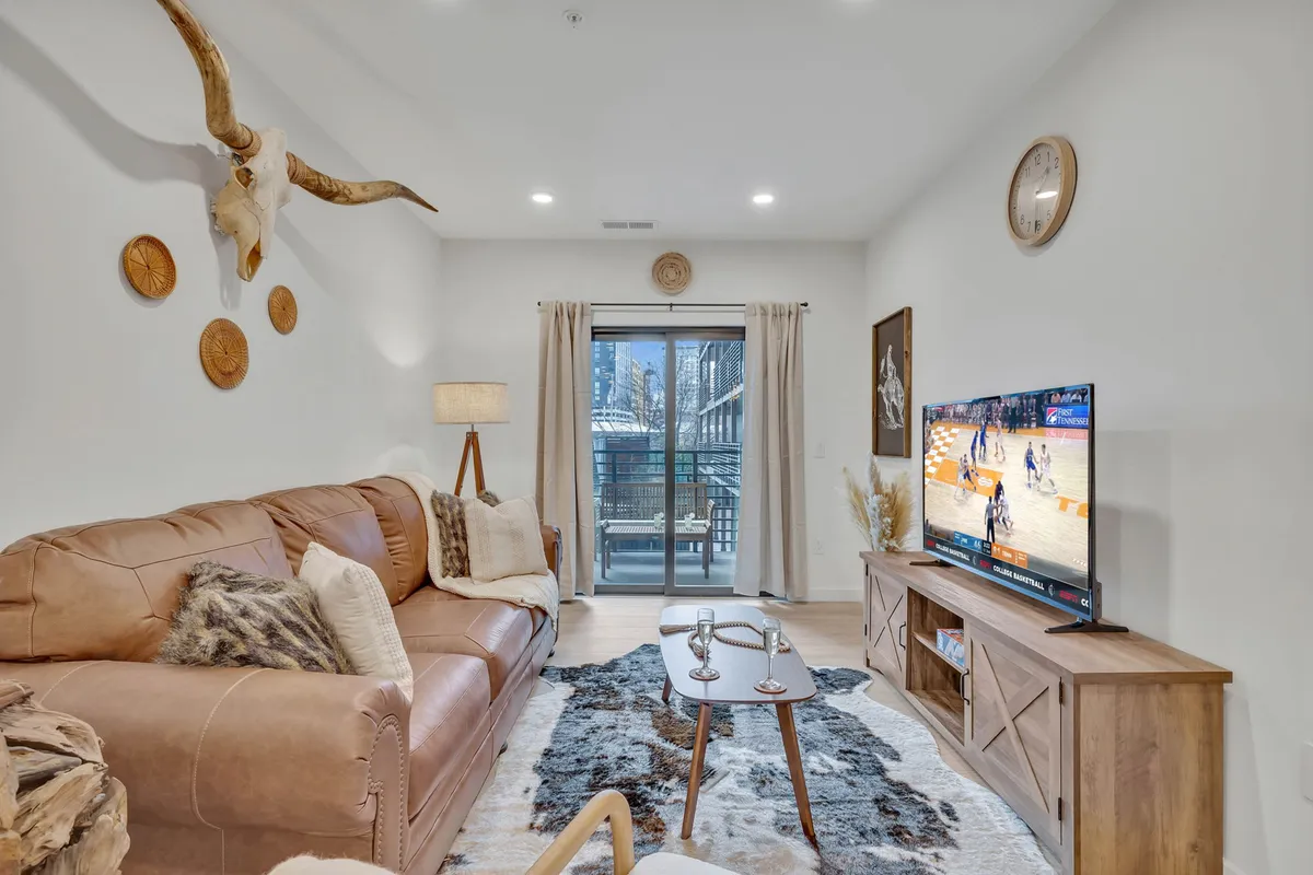 A stylish living room featuring a rich leather sofa, a large television perfect for game days, a striking horn decor piece above, and a rustic wooden coffee table. The space exudes a cozy yet modern Nashville vibe, ideal for gathering, relaxing, and entertaining.