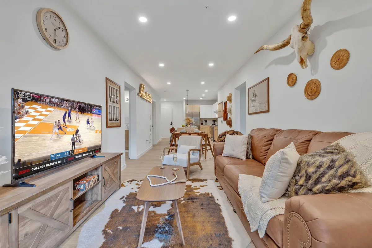 A sunlit, open-concept living area featuring a leather couch, a smart TV displaying a basketball game, decorative wall art, and a dining area in the background. The arrangement encourages socializing, relaxation, and group activities.
