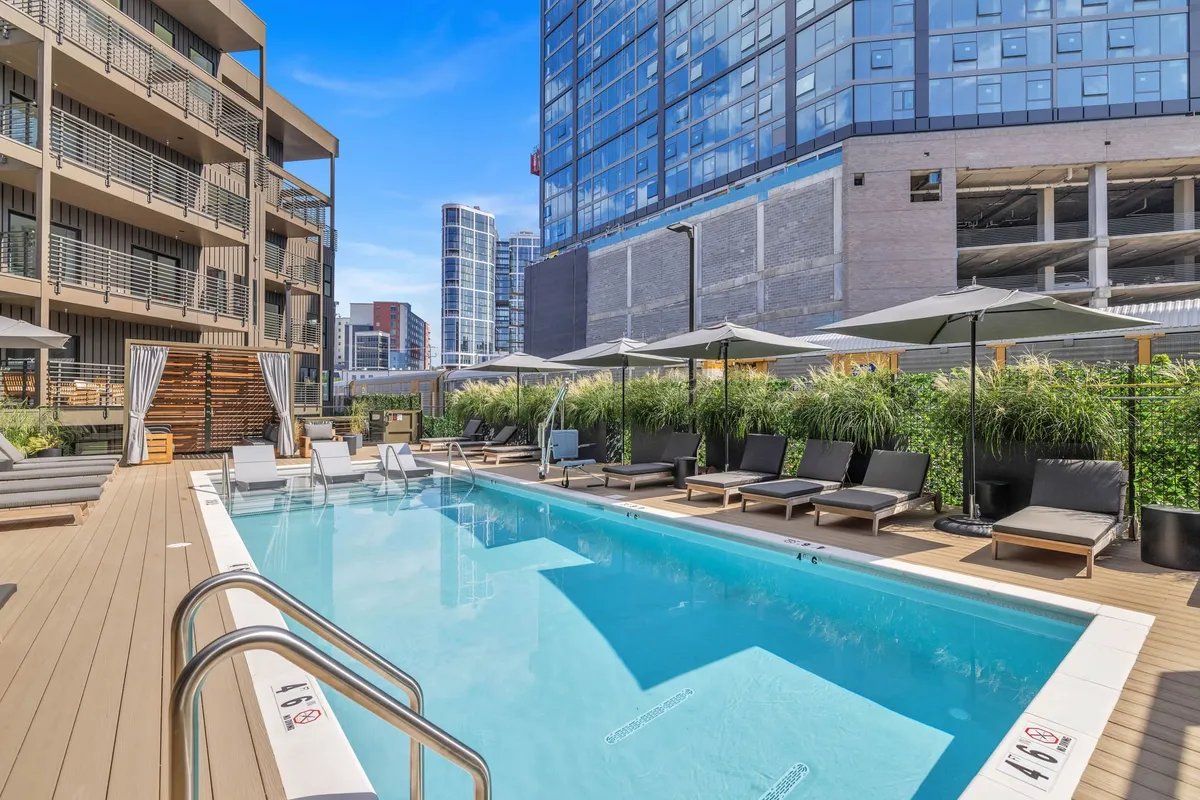 A vibrant exterior scene of a stylish swimming pool surrounded by lounge chairs, umbrellas, lush greenery, and modern buildings in the background. The setup suggests a lively and inviting communal area perfect for swimming, sunbathing, relaxing, and socializing, with hints of amenities like outdoor dining.